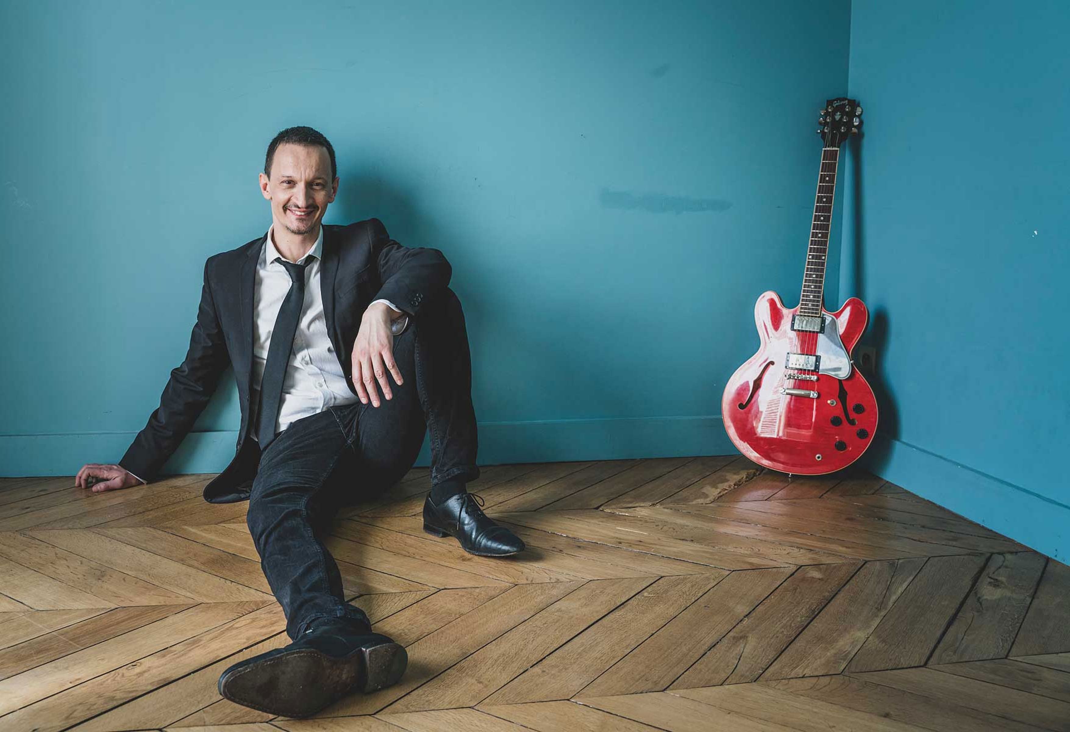 Guillaume Farley, assis au sol à côté de sa guitare, dans un décor bleu-canard